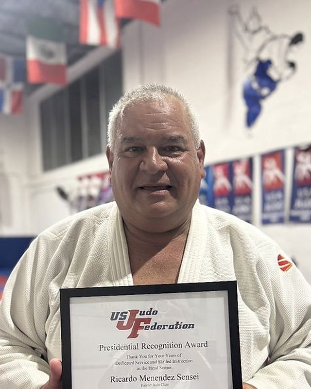 Sensei Ricardo Menendez smiling in his judogi at the dojo, holding a framed certificate.