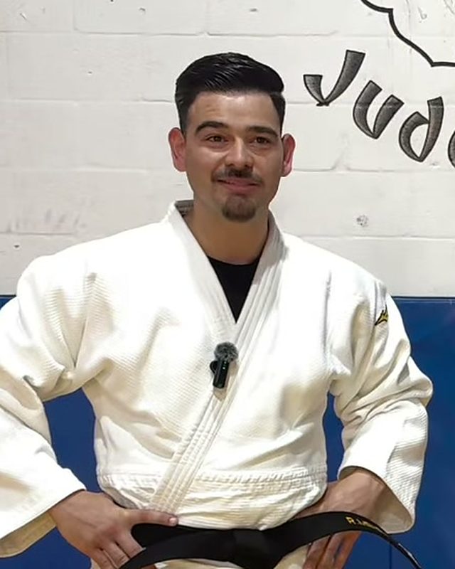 Sensei Ricky Menendez smiling in his white judogi in front of the dojo mural.