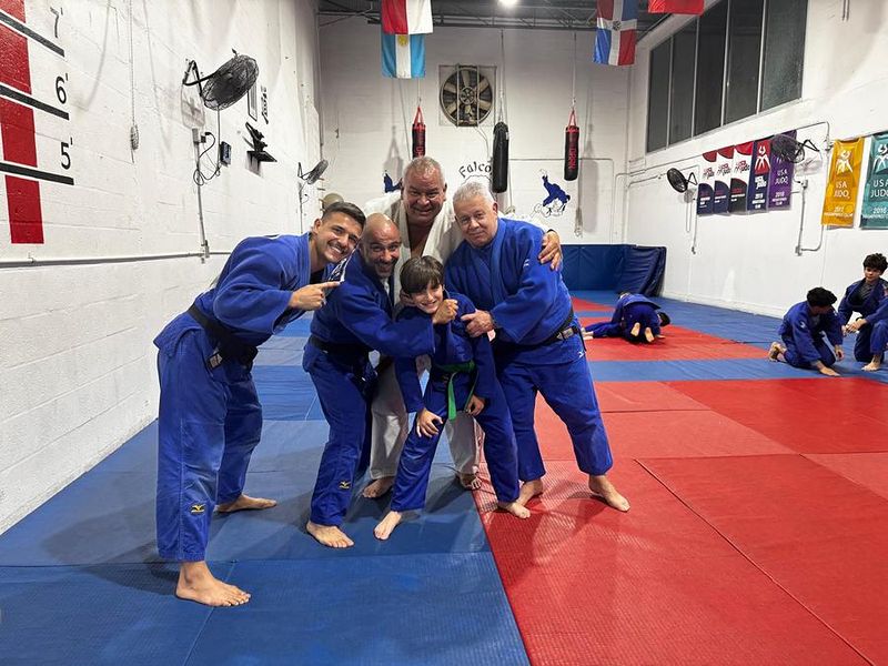 Adult judokas in blue judogis joking around with a young student while others train behind them.