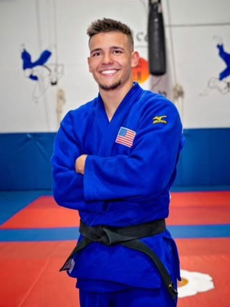 Sensei Alejandro Menendez smiling with his arms crossed, in a blue judogi with a black belt, on the dojo mat.