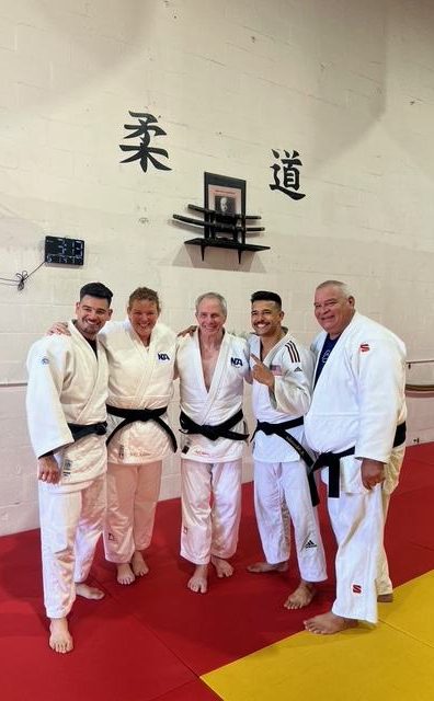 Five black belts in white judogis standing together under the judo characters on the dojo wall.