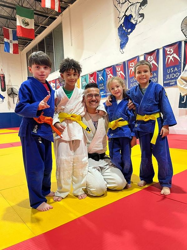 Coach Alejandro Menendez kneeling on the mat with four young students.