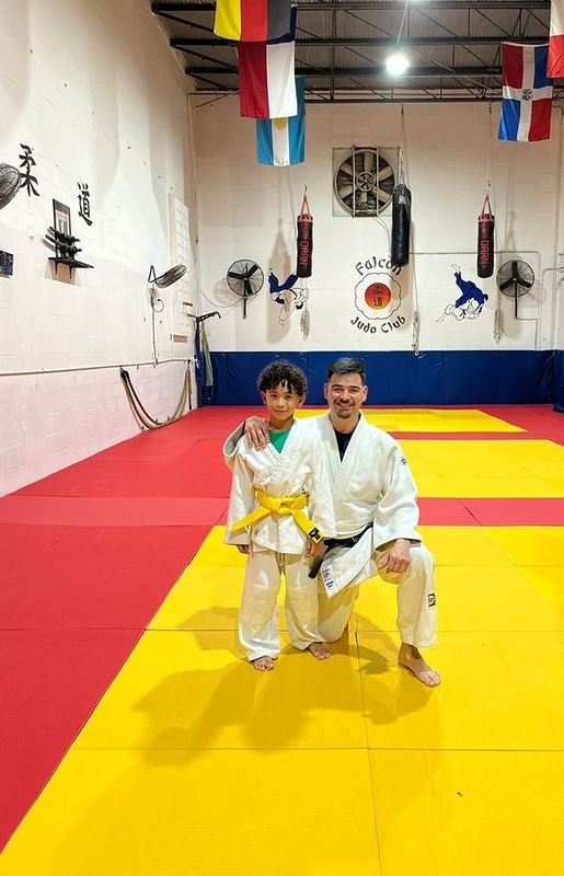 A coach in a white judogi kneeling next to a young student with a yellow belt.